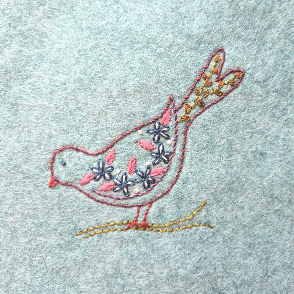 embroidered folk style bird in a branch on a light blue piece of felt.