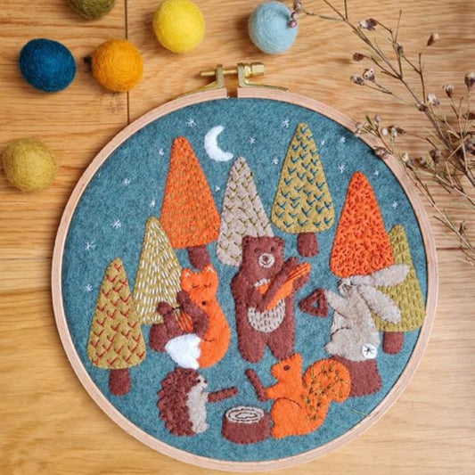 Embroidery hoop with a felt and embroidered design featuring a woodland animals playing music and some trees.  The hoop rests on a  wooden background with a bunch of dried flowers and felt balls.