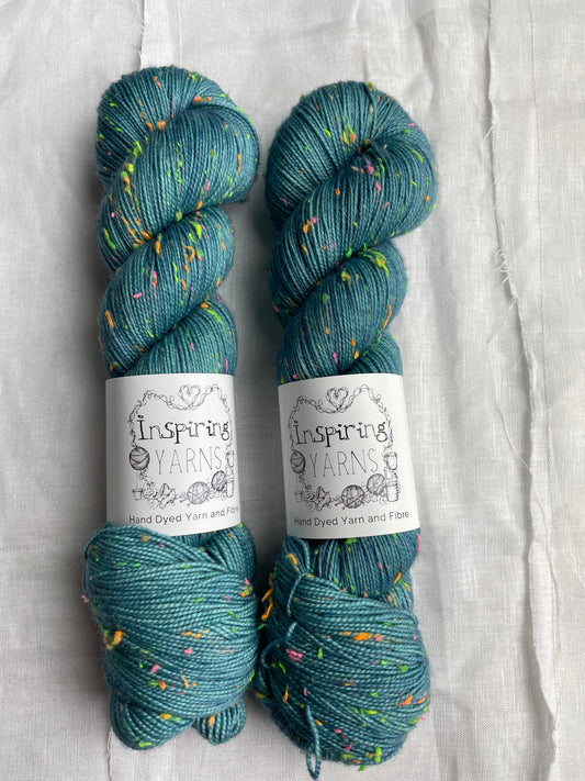 Two skeins of tonal soft teal coloured hand dyed Disco Donegal yarn branded Inspiring Yarns on a white linen background.  The yarn has multi coloured neon neps.