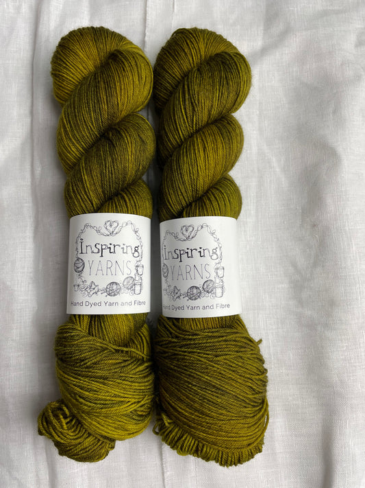 Two skeins of Gremlin, a dark olive green coloured Merino Smooth Sock, labelled Inspiring Yarns on a white linen background. 