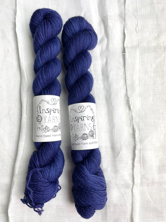 Two tonal deep blue purple coloured skeins of hand dyed Merino Smooth Sock yarn branded Inspiring Yarns on white linen fabric.