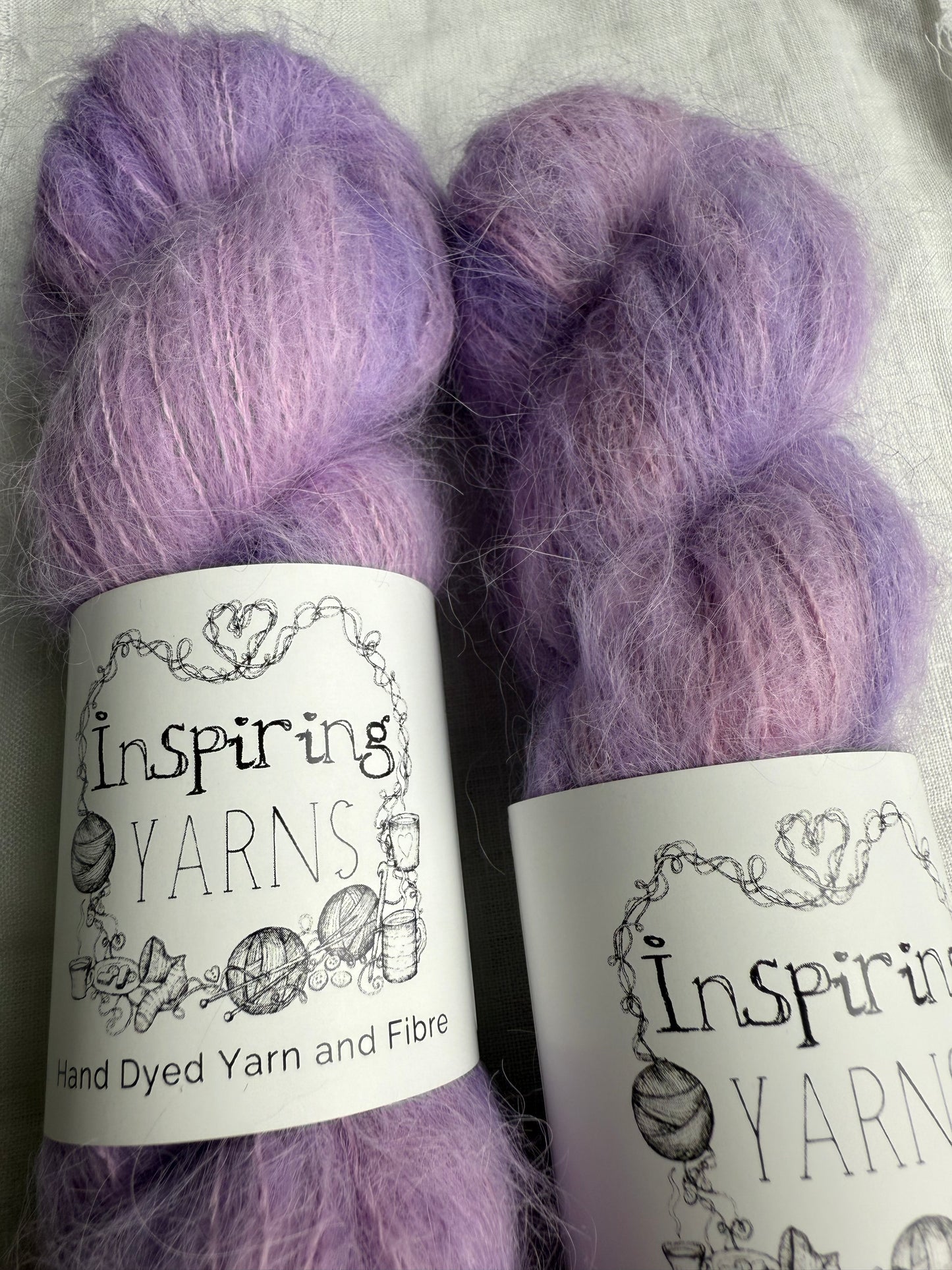 two skeins of light pink and lilac yarn up-close with inspiring yarns labels and white background.