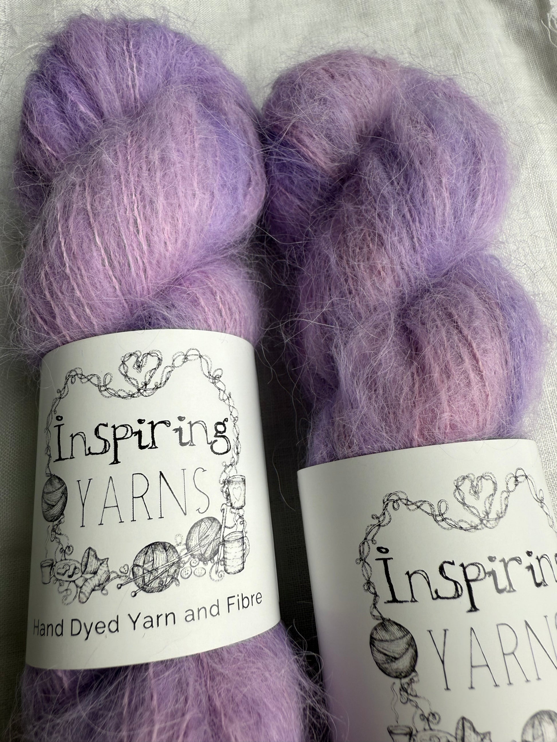 two skeins of light pink and lilac yarn up-close with inspiring yarns labels and white background.