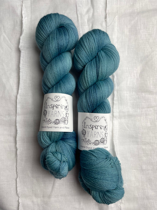 Two skeins of tonal soft teal coloured hand dyed Merino Lace yarn branded Inspiring Yarns on a white linen cloth.