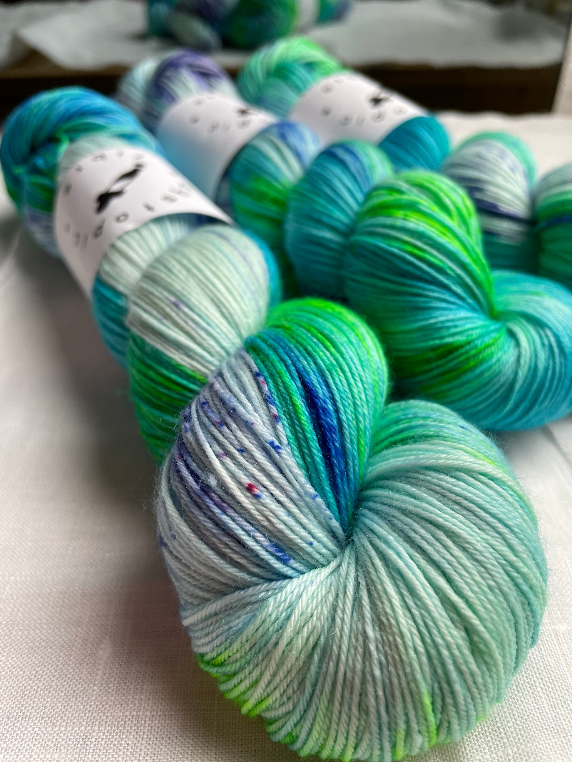 Close up of  3 skeins of turquoise and green hand dyed yarn by Dystopic Fibre on a white table top