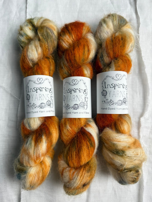Three skeins of fluffy yarn with oranges greys and speckles of different green tones with inspiring yarns label on white background.