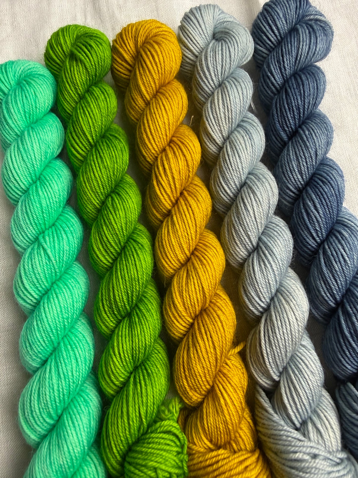 Five mini skeins of yarn in blue, blue grey, yellow, green & bright mint green on a light background