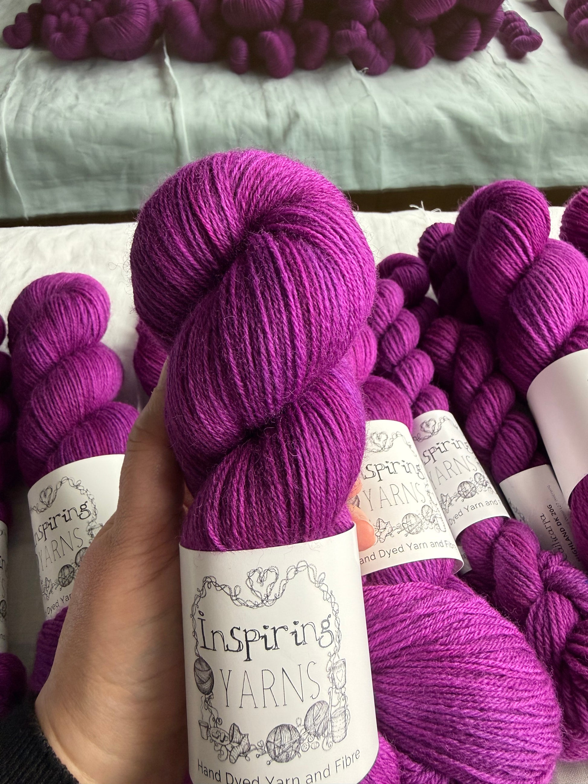 Hand holding a skein  of fuchsia tonal yarn labelled Inspiring Yarns.  More skeins of yarn of the same colour sit in the background.