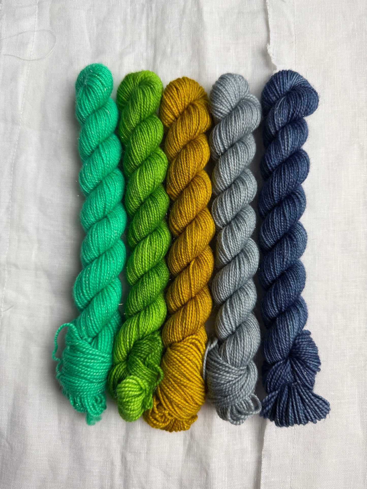 Five mini skeins of yarn in blue, blue grey, yellow, green & bright mint green on a light background