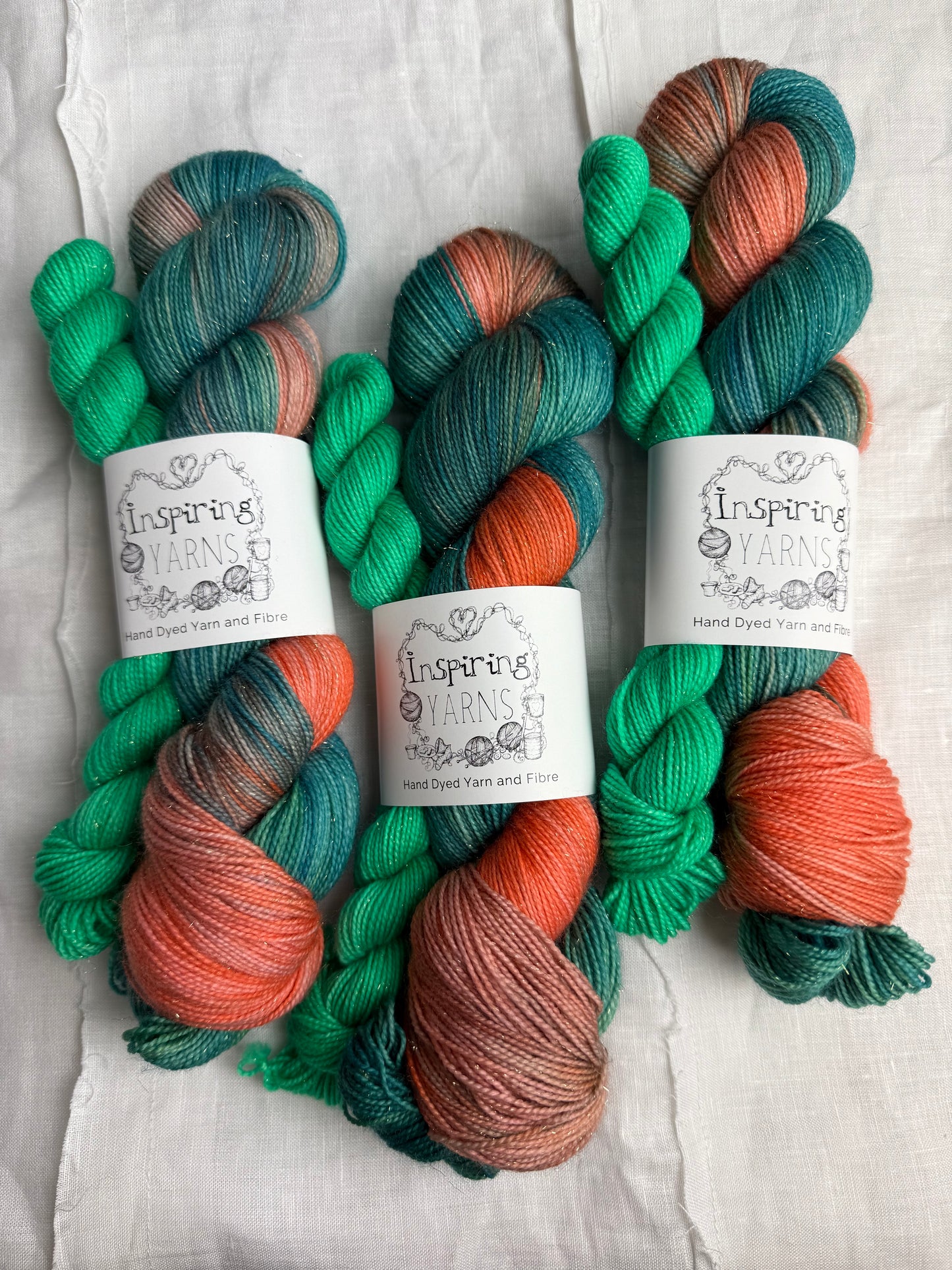 three skeins of hand dyed yarn in greens, peach and teal  on a fabric background and with inspiring yarns labels.