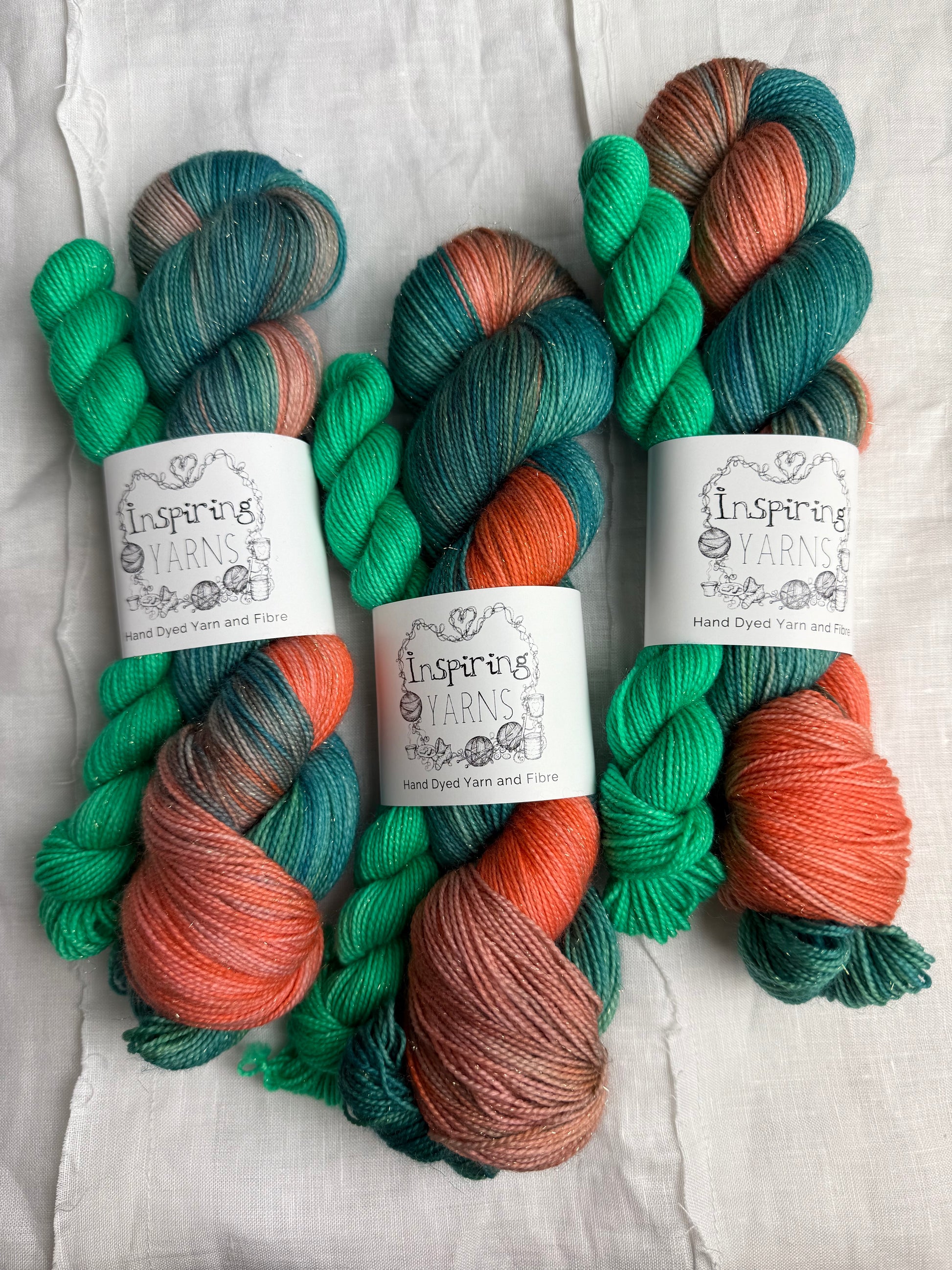 three skeins of hand dyed yarn in greens, peach and teal  on a fabric background and with inspiring yarns labels.