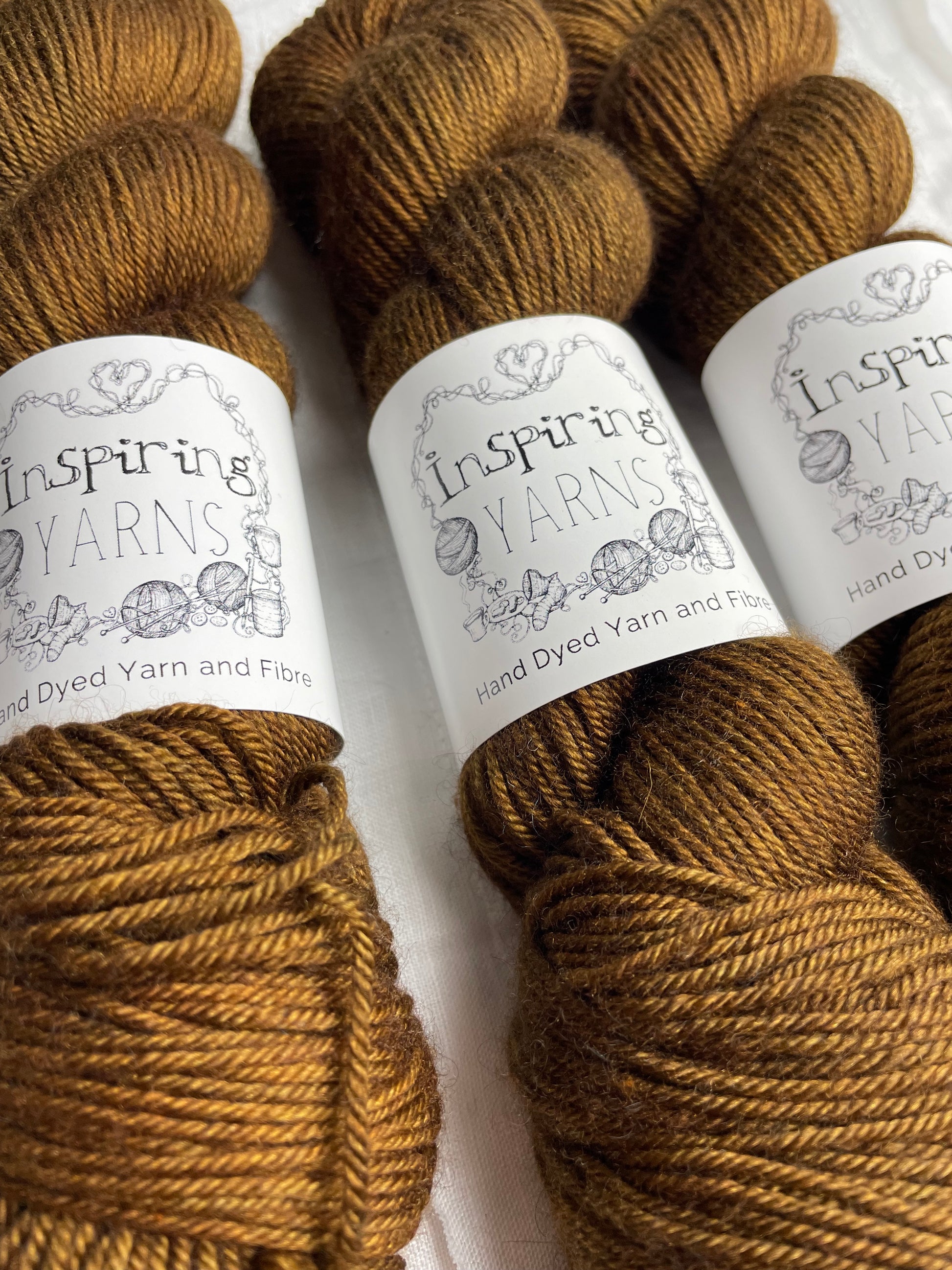 Close up of Three skeins of bronze brown coloured yak silk dk yarn branded Inspiring Yarns on a white linen background.