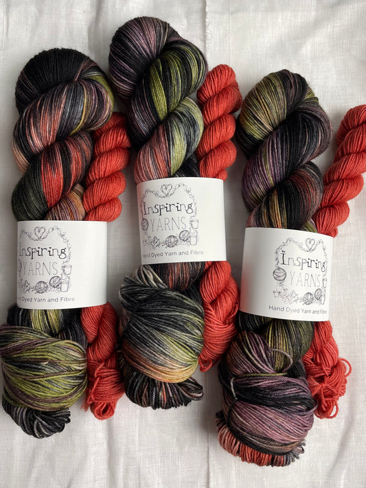 three sock sets made of a skein of black and multi coloured hand dyed yarn with a contrasting deep terraccotta mini skein, labelled Inspiring Yarns and laid on a white linen cloth.