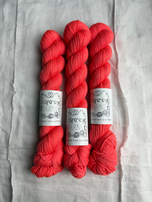 three skeins of deep neon coral yarn on a white background and inspiring yarns label around them.