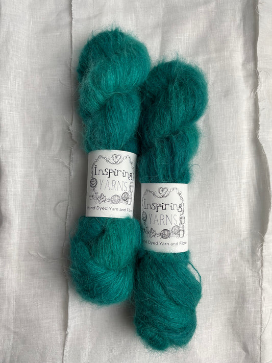 Two skeins of fluffy brushed suri silk lace yarn hand dyed in a tonal jade green colourway and labelled Inspiring Yarns on a white linen background.