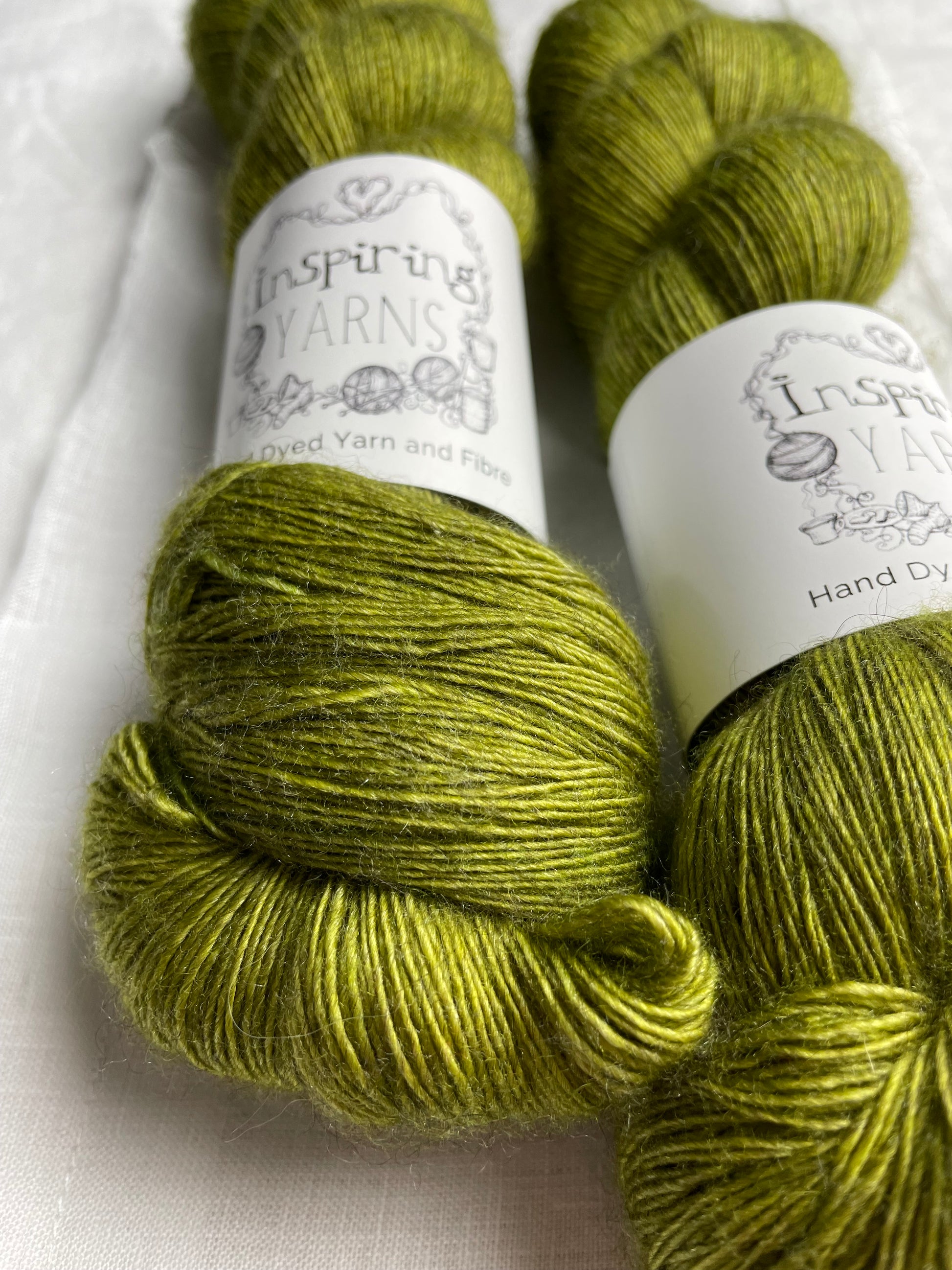 Close up of two tonal spring green coloured skeins of hand dyed Yak Silk Singles branded Inspiring Yarns on a white linen background.