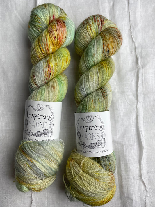 Two skeins of  speckled silver grey and mint coloured hand dyed Merino Lace yarn branded Inspiring Yarns on a white linen cloth.