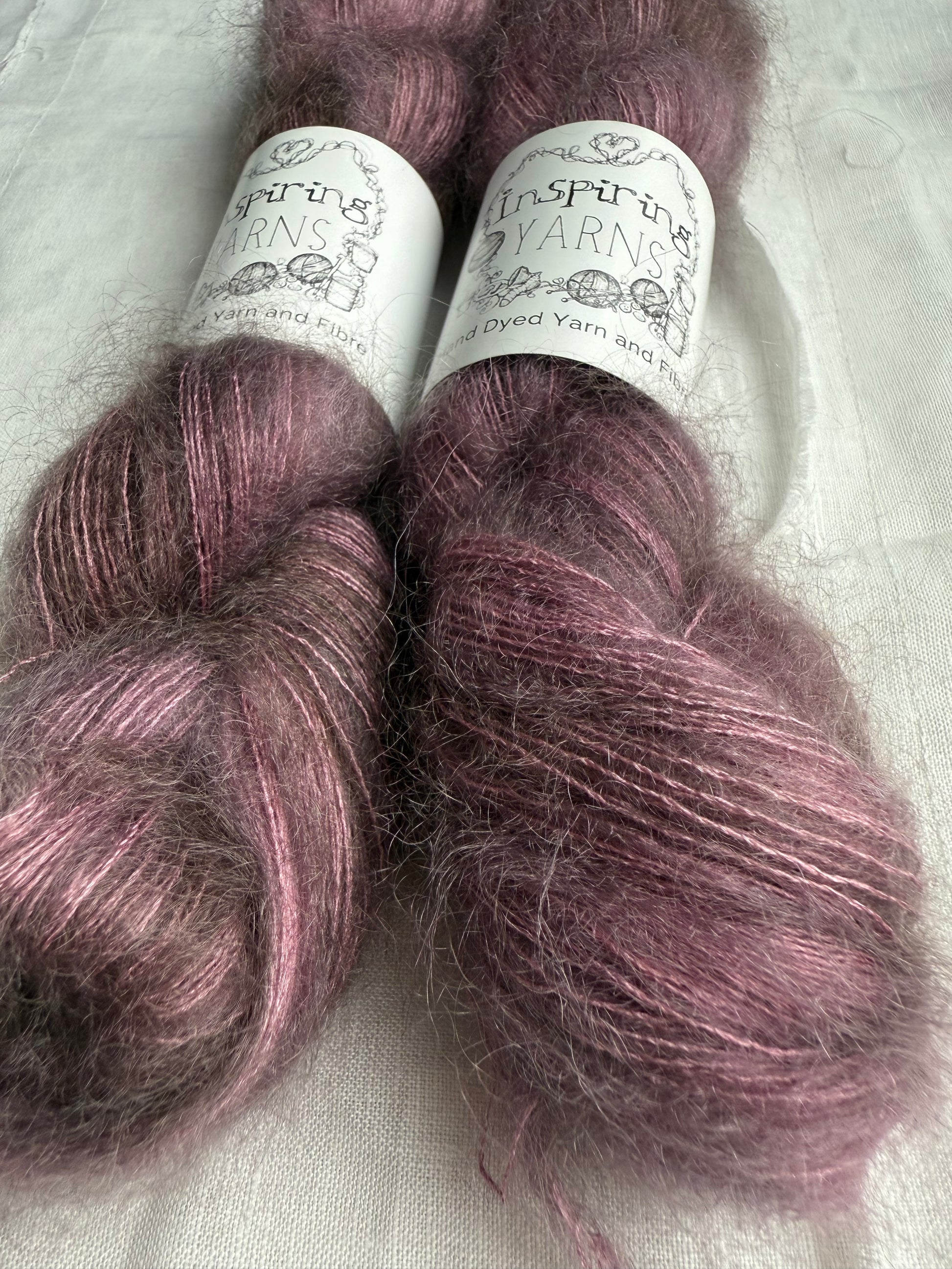 Two skeins of dusty purple yarn with labels on a white fabric background