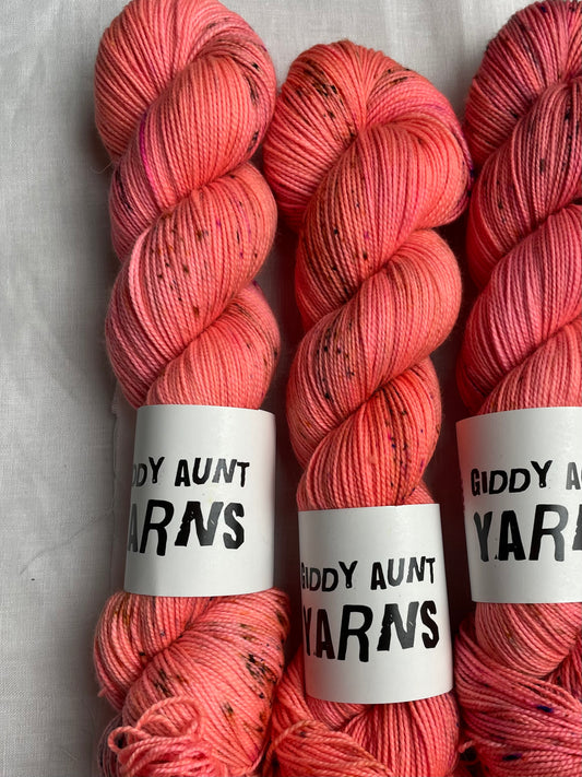 3 skeins of hand dyed yarn in a bright neon coral colour with dark speckles on a white background, the yarn band reads giddy aunt yarns