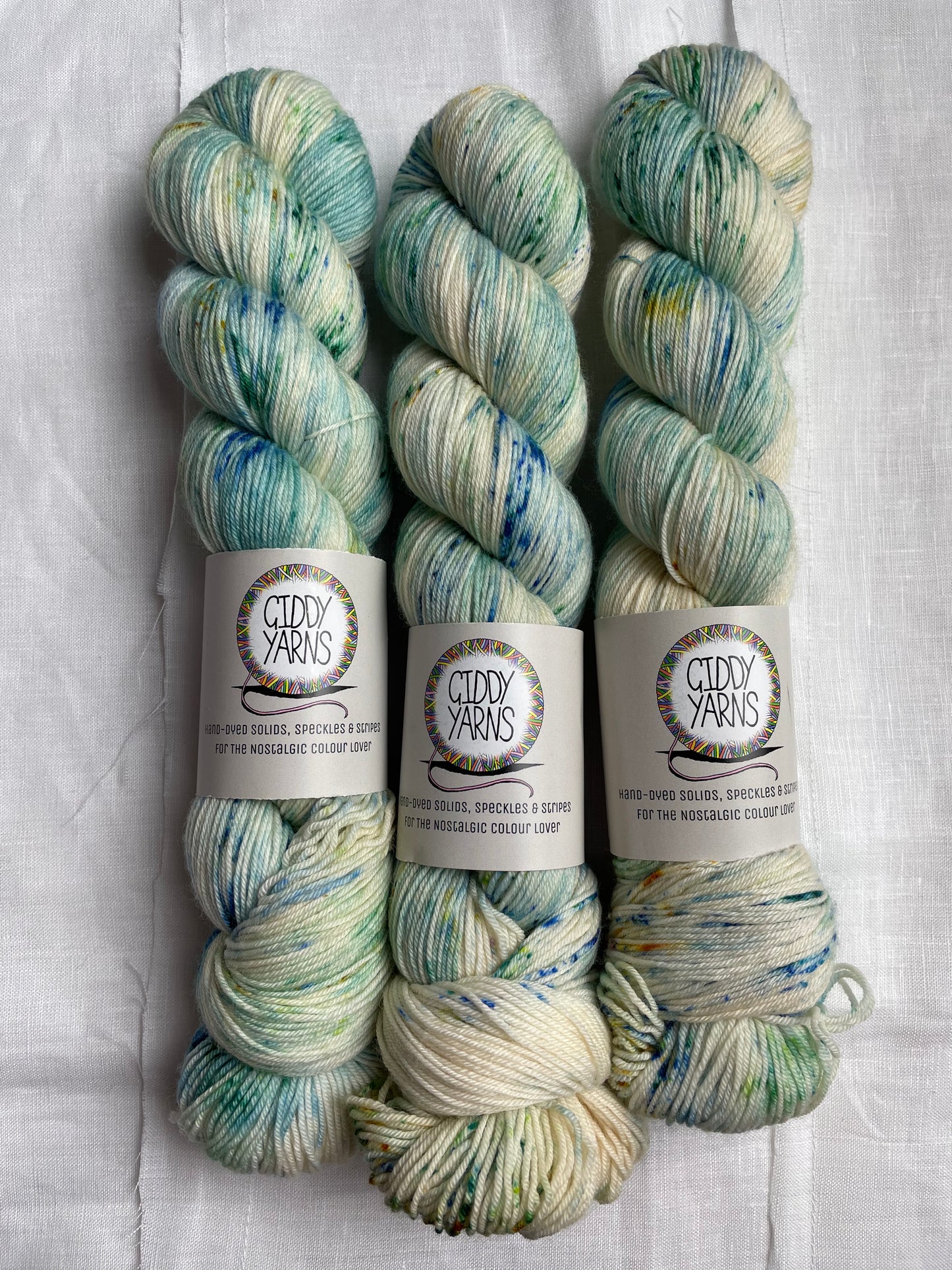 3 skeins of hand dyed yarn speckled with shades of blue green and teal, the yarn bad reads Giddy Yarns, on a white background