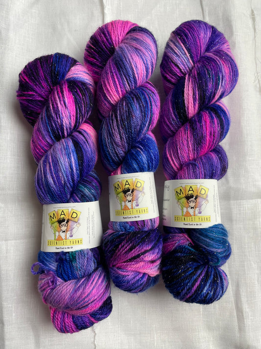 3 skeins of hand dyed yarn in shades of bright pink, purple and blue with silver lurex sparkles lie on a white background, the yarn band reads mad scientist yarns