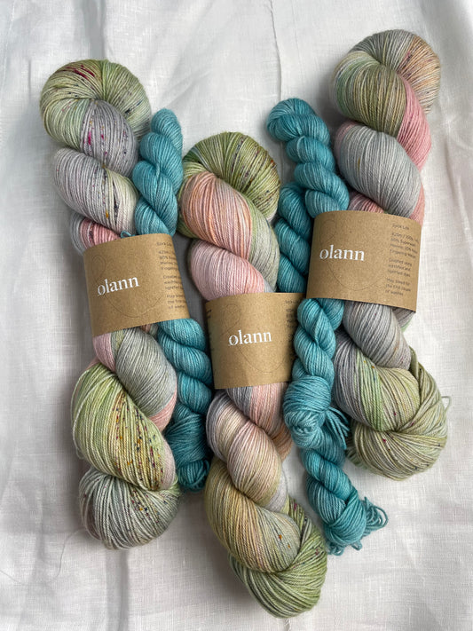 3 sock sets of hand dyed yarn , the main skein is multi coloured pastel shades and it is paired with a light turquoise mini skein, the yarn is labelled Olann