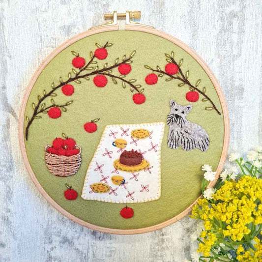 Embroidery hoop with a felt and embroidered design featuring a dog, apples, a laid out picnic and basket design.  The hoop rests on a white wooden background with a bunch of yellow flowers.