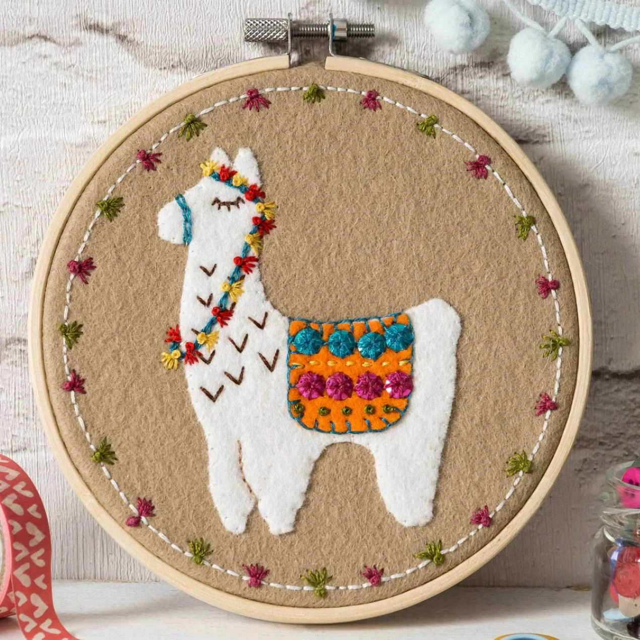 Embroidery hoop with a felt and embroidered design featuring a white llama in a patterned rug.  The hoop rests on a white wooden background.