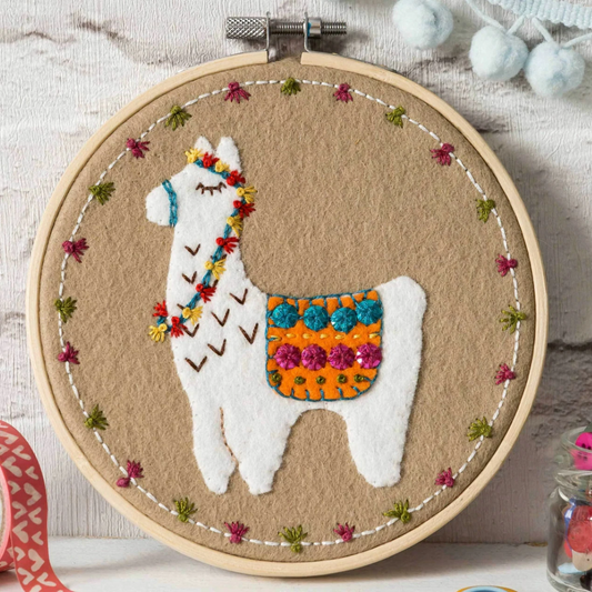 Embroidery hoop with a felt and embroidered design featuring a white llama in a patterned rug.  The hoop rests on a white wooden background.
