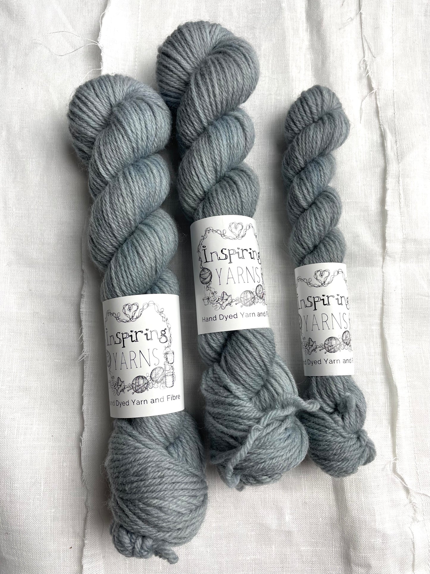 Three skeins of a tonal light grey blue Highland DK yarn branded Inspiring Yarns on a white linen background.