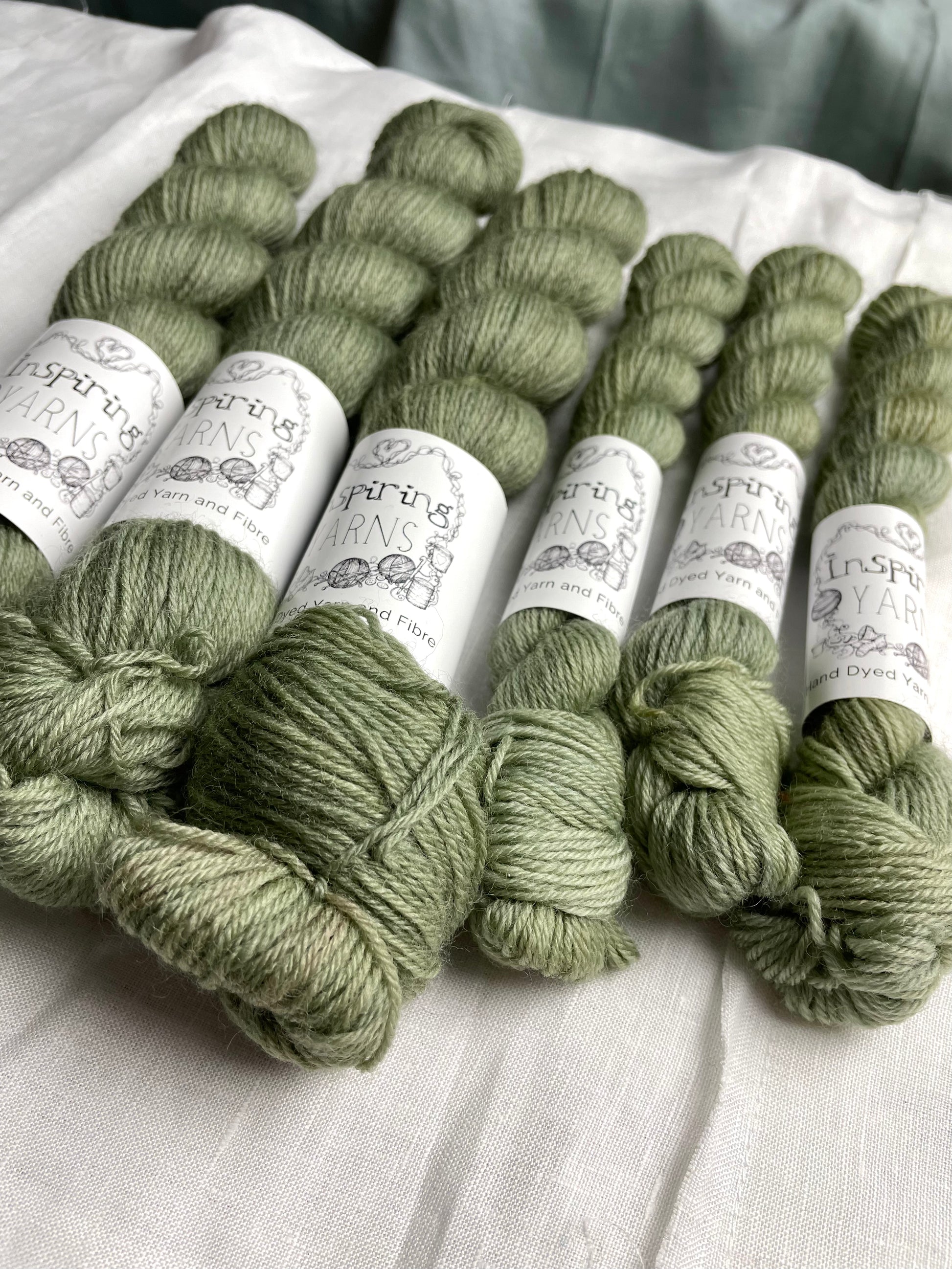 Close up of some skeins of tonal light green grey coloured Highland 4Ply yarn branded Inspiring Yarns on a white linen background.