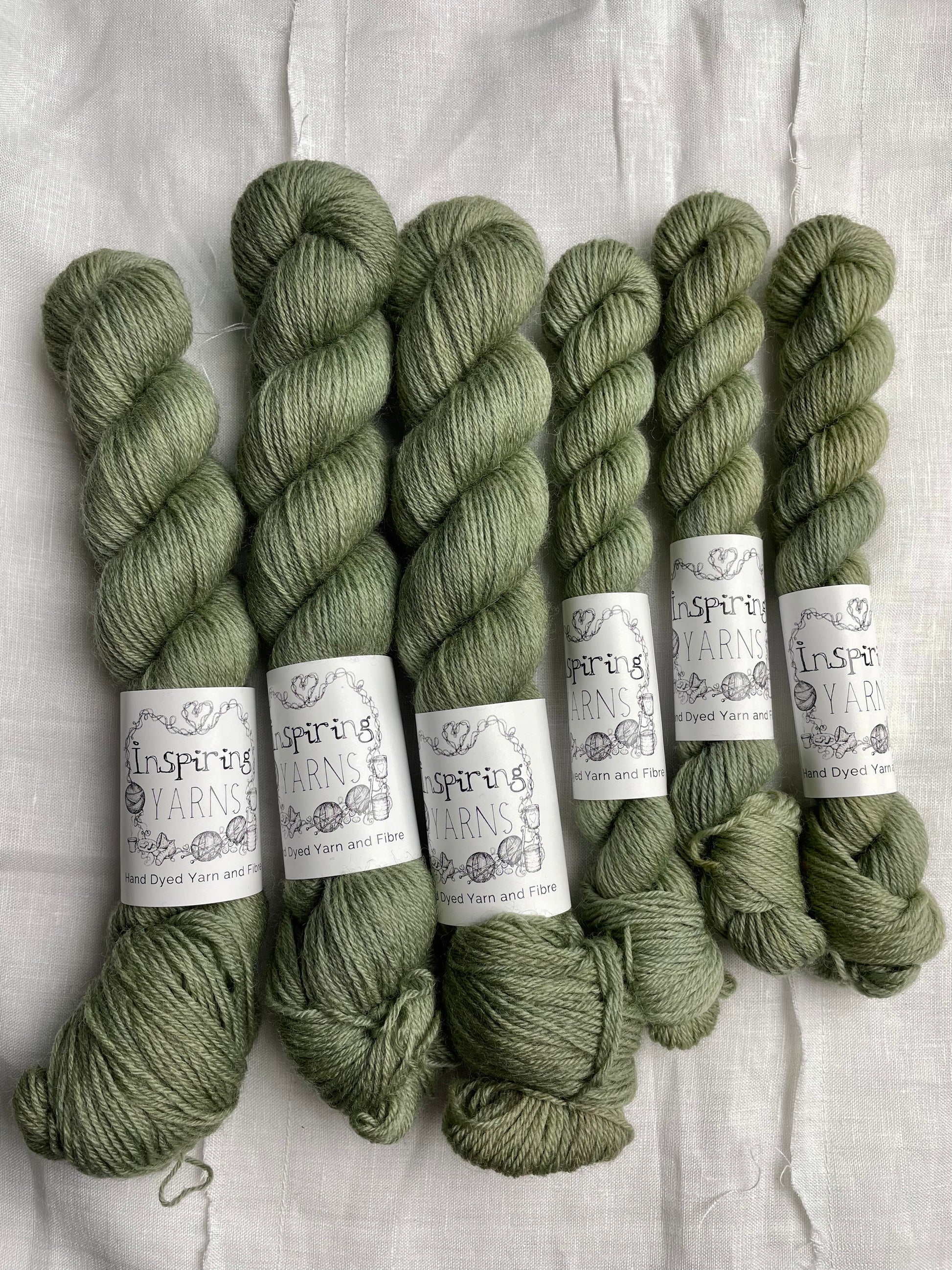 Skeins of tonal light green grey coloured Highland 4Ply yarn branded Inspiring Yarns on a white linen background.