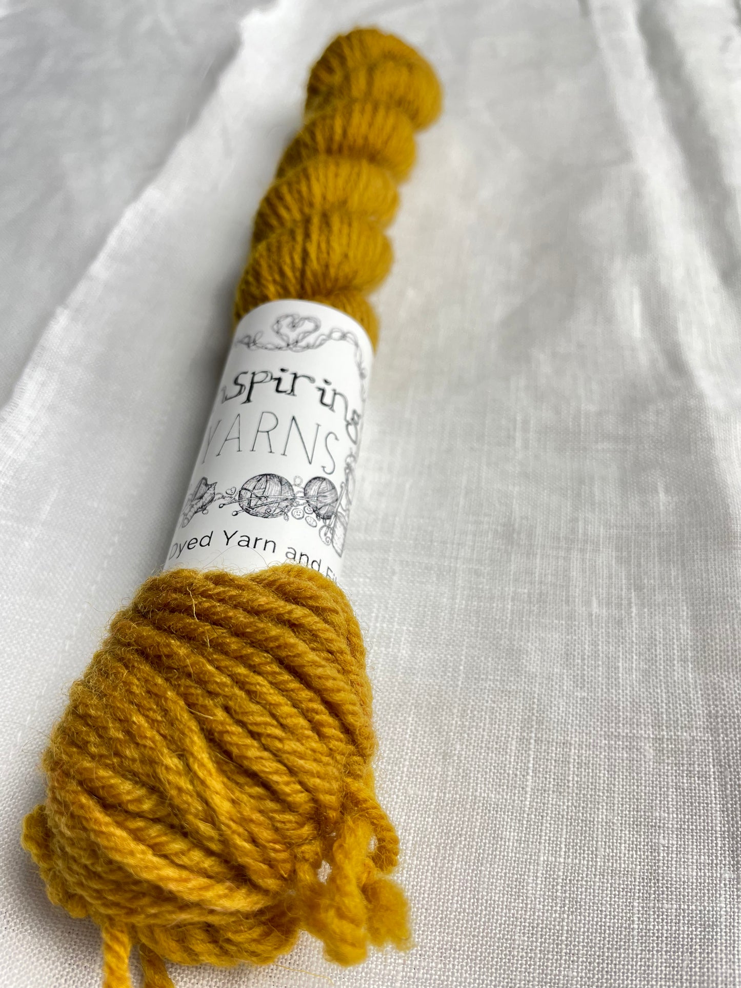 Close up of one skein of a tonal mustard coloured mini skein of Highland DK on a white linen background.