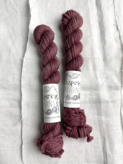 Two mini skeins of a tonal dark mauve Highland DK yarn branded Inspiring Yarns on a white linen background.