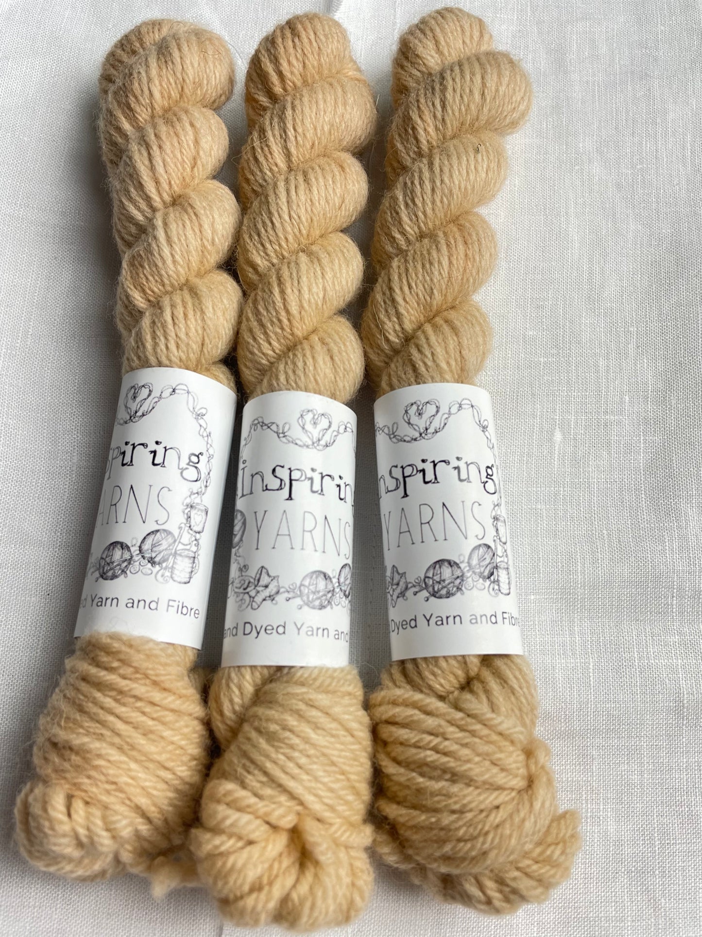 Three mini skeins of soft golden cream coloured Highland DK yarn branded Inspiring Yarns on a white linen background.