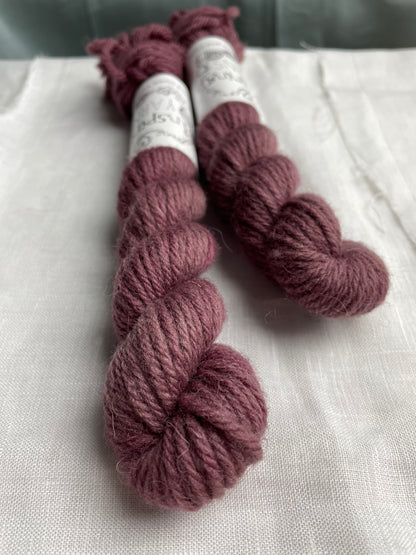 Close up of two mini skeins of a tonal dark mauve Highland DK yarn branded Inspiring Yarns on a white linen background.