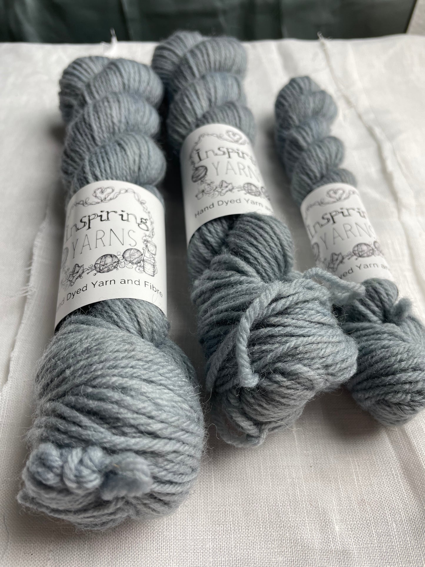 Close up of three skeins of a tonal light grey blue Highland DK yarn branded Inspiring Yarns on a white linen background.