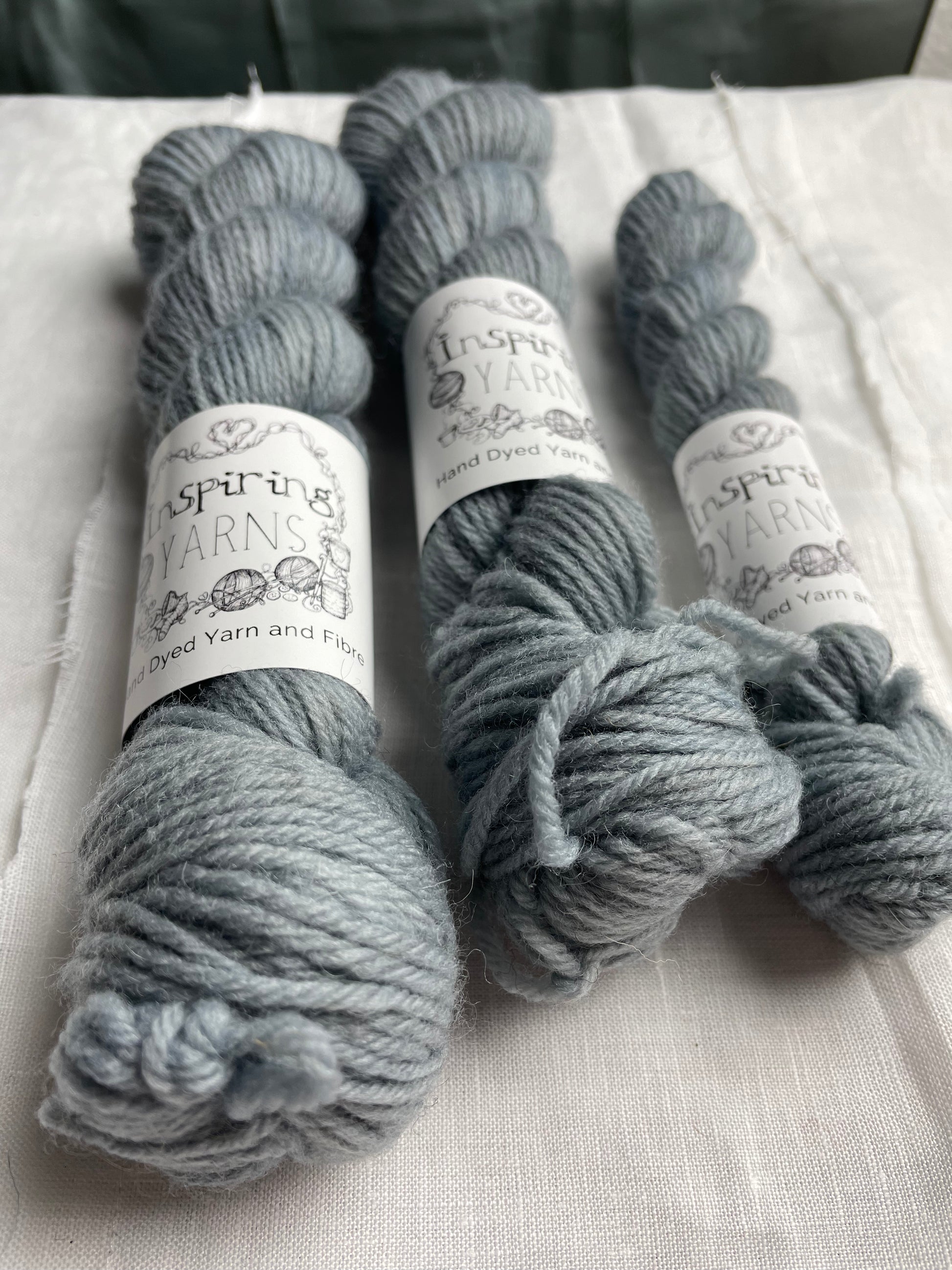 Close up of three skeins of a tonal light grey blue Highland DK yarn branded Inspiring Yarns on a white linen background.