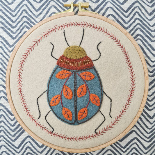 Embroidery hoop with a felt and embroidered design featuring beetle with a leaf design on its back.  The hoop rests on blue striped fabric background.