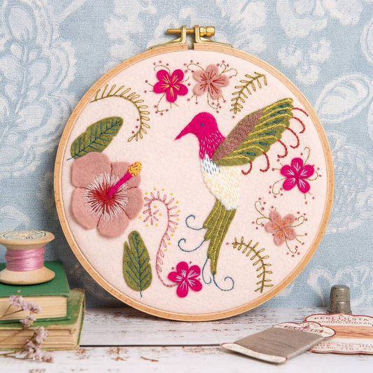 Embroidery hoop with a felt and embroidered design featuring a hummingbird and flowers.  The hoop rests on a blue background with some thread, dried flowers, books and a thimble in the foreground.