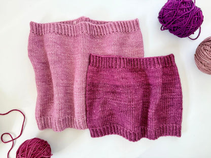 Two knitted cowls in different shades of purple. with a number of balls of yarn on a plain white surface.